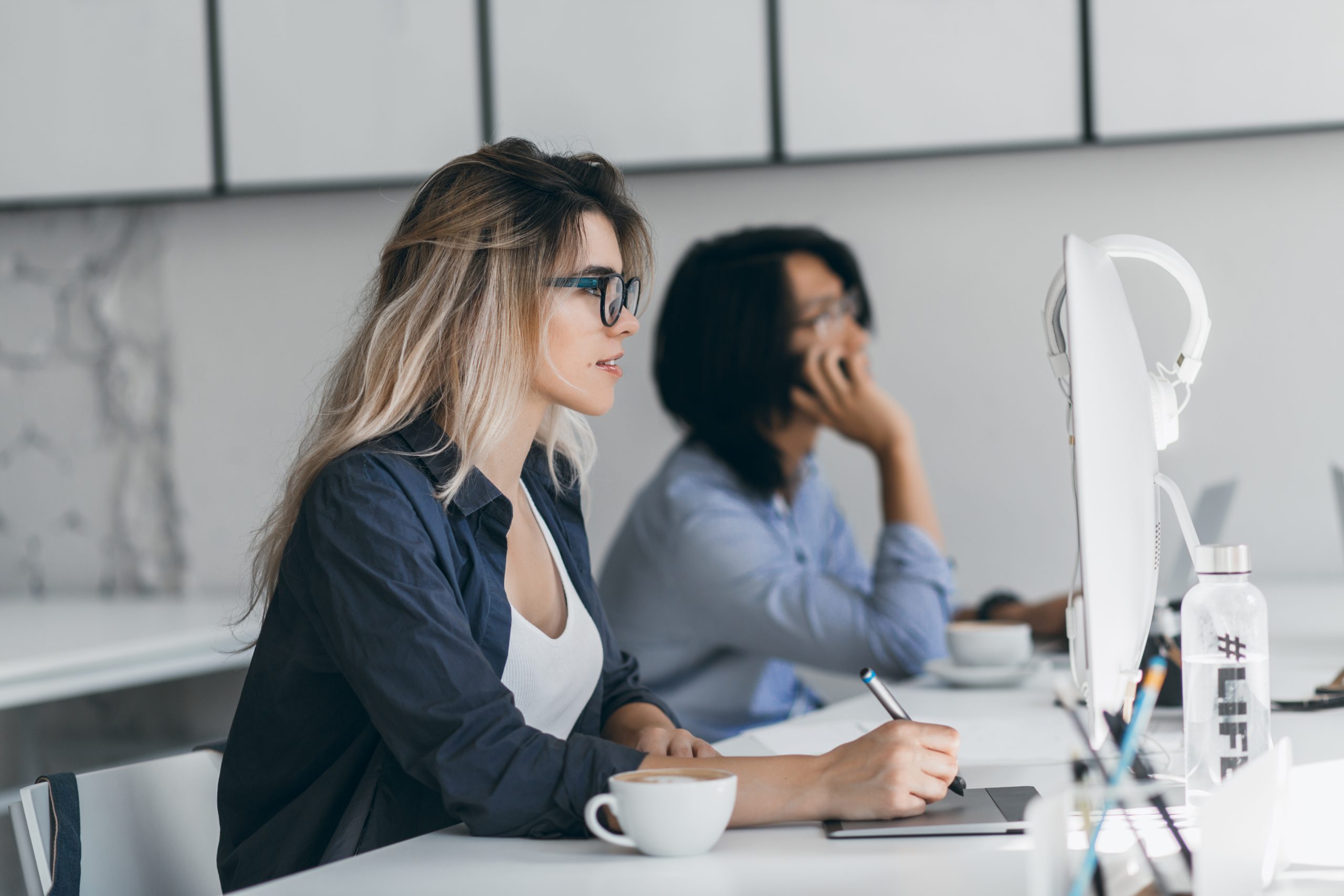 Inspired freelance web-designer using tablet and stylus, looking at screen while her friend talking on phone. Asian student holding smartphone and typing on keyboard, sitting beside blonde girl..